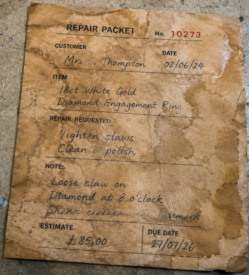 A heavily water-damaged brown jewellery repair packet on a workshop bench, with only the repair number and a few scattered words still clearly readable